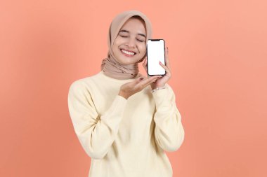 Heyecanlı genç Asyalı Müslüman kadın tesettür ve süveter giyiyor. Akıllı telefonu boş ekran, boş ekran ile gösteriyor. Şeftali stüdyosunun arka grubunda izole edilmiş uygulamayı öneriyor.