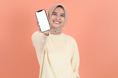 Heyecanlı genç Asyalı Müslüman kadın tesettür ve süveter giyiyor. Akıllı telefonu boş ekran, boş ekran ile gösteriyor. Şeftali stüdyosunun arka grubunda izole edilmiş uygulamayı öneriyor.