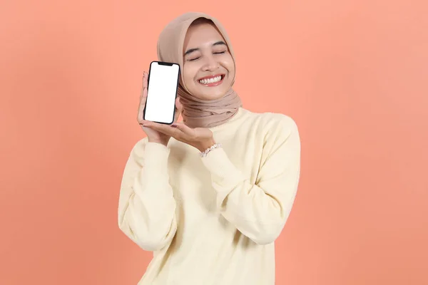 stock image Excited beautiful young Asian Muslim woman wearing hijab and sweater showing smartphone with empty screen, blank screen recommending App isolated on peach studio backgroun