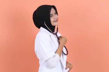 Güzel bayan doktor Asyalı Müslüman tesettür steteskop kullanıyor ve şeftali arka planına bakıyor.