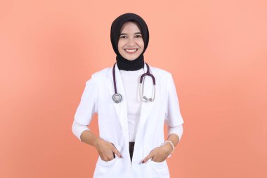 Doktor ceketli güzel bir doktor neşeli bir yüzle duruyor ve ellerini cebine sokuyor şeftali arka planına steteskop asıyor.
