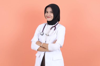 Asyalı Müslüman güzel bayan doktor, beyaz önlüklü, steteskoplu, belini yukarıda. Kadın hastane çalışanı kameraya bakıp gülümsüyor.