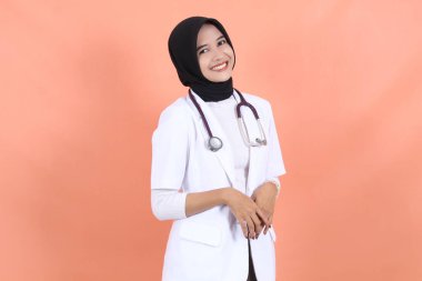 Asyalı Müslüman güzel bayan doktor, beyaz önlüklü, steteskoplu, belini yukarıda. Kadın hastane çalışanı kameraya bakıp gülümsüyor.