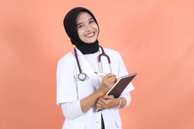 Peçeli, bayan, Asyalı Müslüman doktor. Steteskoplu, beyaz takım elbiseli, beli kalkık ve deftere yazı tarifi yazarak gülümsüyor..