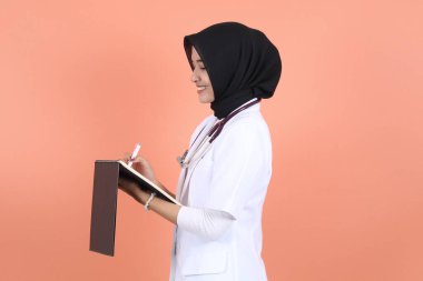 Peçeli, bayan, Asyalı Müslüman doktor. Steteskoplu, beyaz takım elbiseli, beli kalkık ve deftere yazı tarifi yazarak gülümsüyor..