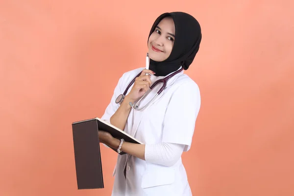 veiled female asian muslim doctor wearing white suit uniform with stethoscope, waist up and smiling with writing recipe on notebook.