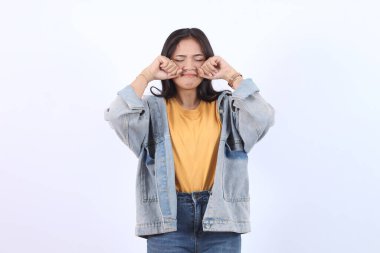 Asyalı kadın iki yanağını tutarken üzgün bir ifadeyle ağlarken mavi ceketli, sarı tişörtlü, izole edilmiş beyaz arka plan.