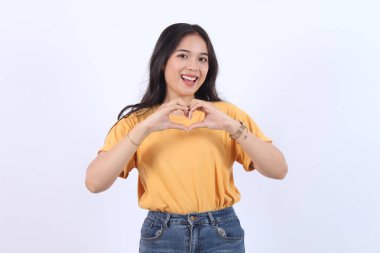 Gülümseyen genç kız iki eliyle kalbini gösteriyor, aşk işareti açık mavi arkaplanda izole edilmiş genç Asyalı kadın el kol hareketleri yapan kalp şeklinde gülümseyen Asyalı kız aşık oluyor