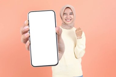 Gülümseyen genç Asyalı Müslüman kadın reklam için cep telefonu ekranı gösteriyor ve şeftali arka planda izole edilmiş başarıyı kutluyor. Halk Yaşam Tarzı Konsepti