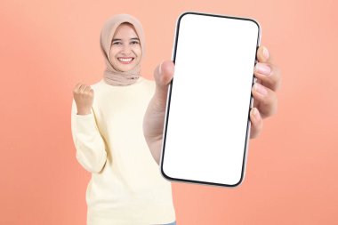 Gülümseyen genç Asyalı Müslüman kadın reklam için cep telefonu ekranı gösteriyor ve şeftali arka planda izole edilmiş başarıyı kutluyor. Halk Yaşam Tarzı Konsepti