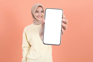 Gülümseyen genç Asyalı Müslüman kadın reklam için cep telefonu ekranı gösteriyor ve kalçalarını şeftali arkaplan üzerinde tutuyor. Halk Yaşam Tarzı Konsepti