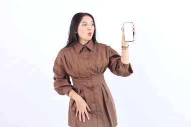 Kahverengi elbiseli, gülümseyen genç Asyalı kadın beyaz arka planda izole edilmiş bir reklam için cep telefonu ekranını gösteriyor. Halk Yaşam Tarzı Konsepti