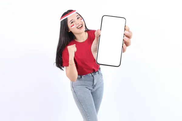 Stock image Excited young Asian woman with red white flag ribbon showing a screen mobile phone, for advertisement and presenting celebrating victory Indonesian independence day 17 Agustus. Merdeka concept