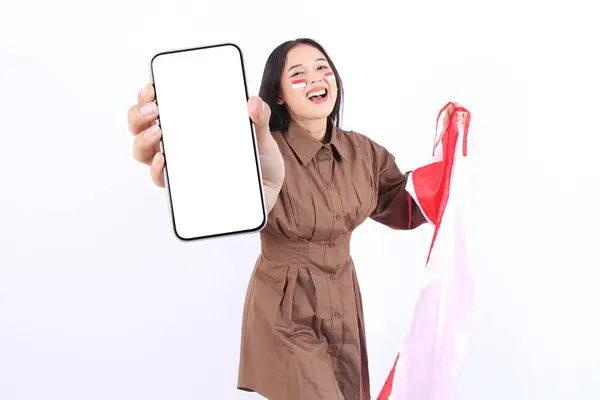 stock image Excited young Asian woman in brown dress showing a screen mobile phone, for advertisement and presenting celebrating victory Indonesian independence day 17 Agustus. Merdeka concept