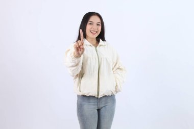 A cheerful adult Asian woman in a casual cream jacket and jeans, smiling while pointing upward with her index finger. showing number 1 with finger Perfect for concepts like ideas, attention clipart