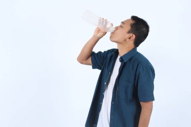 Yetişkin yakışıklı Asyalı adam şişeden su içiyor. Şişe beyaz ve adam mavi gömlek giyiyor. Susamış, beyaz tenli.
