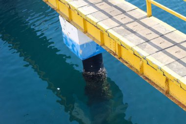 Rıhtımda mavi ve beyaz şamandıralı bir su üzerinde köprü. Su sakin ve köprü sarı.