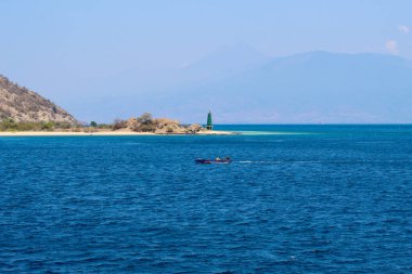 Tekne yeşil bir deniz fenerinin yanında okyanusta yüzüyor. Su sakin ve gökyüzü berrak.