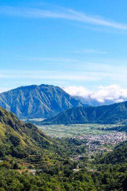 Sembalun Tepesi, Lombok Endonezya sıradağları ve aşağıdaki vadide bir şehir. Gökyüzü açık ve mavi. Dağlar ağaçlarla kaplı ve vadi yemyeşil bitkilerle dolu.