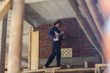 Asyalı işçi, kırmızı kil tuğlayı metal bir malayla duvara sığdırıyor. Kırsal bir inşaat alanında geleneksel ev inşa süreci. 6 Ağustos 2025, Lombok, Endonezya.