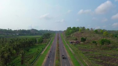 Üzerinde birkaç araba ve arka planda birkaç ağaç olan bir yol. Yol boş ve gökyüzü temiz.