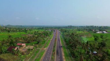 Arka planda bir sürü ağaç ve ev olan bir yol. Birkaç araba ve motosikletin geçtiği yol.
