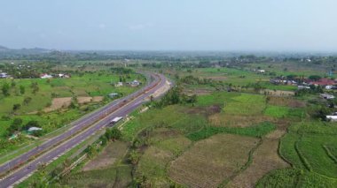 Arka planda bir sürü yeşil çimen ve ağaç olan bir yol. Birkaç araba ve motosikletin geçtiği yol.
