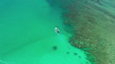 Sörfçüleri ve iki sörf tahtasını taşıyan bir teknenin sinematik görüntüsü. Yeşil deniz ışıldıyor, mercan resifleri sağda görünüyor ve kamera çarpıcı sahnenin üzerinde düzgün dönüyor..
