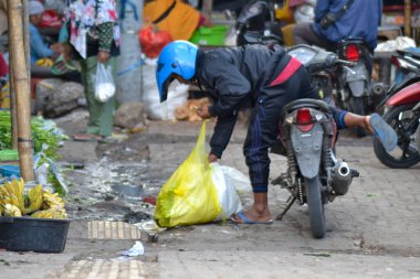 Kasklı bir adam, kalabalık bir markette motosikletinin yanında plastik bir alışveriş çantası taşır..