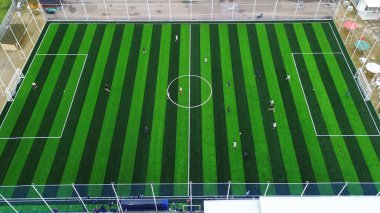 Top-down drone shot of a football game on a striped green artificial turf pitch. Athletes compete in a local league, showing teamwork and strategy.