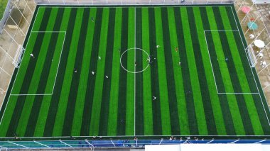 Top-down drone shot of a community football game. Players compete on a vibrant green striped pitch, showcasing teamwork, strategy, and recreation.