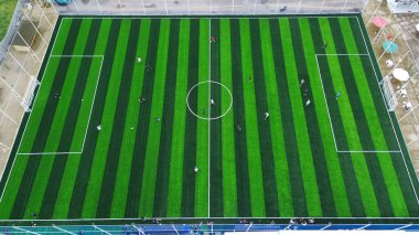 Top-down drone shot of a community soccer game. Teams compete on a striped green artificial turf field, showing teamwork and healthy recreation.