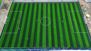 Top-down drone shot of a green striped football pitch. Players compete in a local game, embodying teamwork, strategy, and an active lifestyle.
