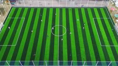 High angle drone shot of a local soccer game. Modern artificial turf with a striped pattern contrasts with the surrounding rural green landscape.