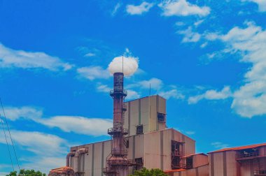 Endüstriyel bacanın mavi gökyüzüne beyaz duman salması üretim faaliyetlerini ve çevresel sorunları sembolize ediyor. Sahne endüstri ve doğanın kesişimlerini yakalıyor.