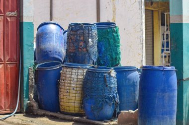 Kırsal bir duvara dayanan renkli plastik variller, kentsel depolama çözümleri ve geri dönüşüm uygulamalarını gösteriyor.