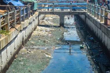 Kentsel su yolu çöplerle tıkanmış, çevre kirliliği sorunlarını vurguluyor. Plastik şişeler ve torbalar molozların arasında yüzer ve atıkların uygun olmayan bir şekilde atılmasının etkisini gösterir.