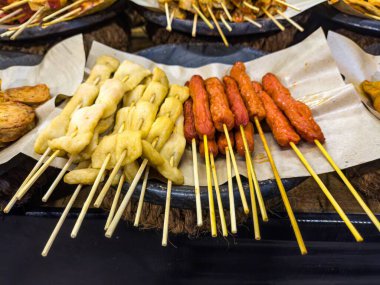 A variety of skewered street foods, including fish cakes and sausages, displayed on parchment paper at a traditional angkringan stall clipart