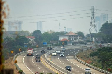 Arka planda yeşillik ve kentsel binalarla çevrili, birden fazla aracı olan işlek bir otoyol. Kentsel altyapı ve ulaşım temaları için ideal