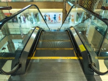 Bir alışveriş merkezinde modern bir yürüyen merdiven. Temiz cam kenarları ve parlak bir tasarımı var. Müşterilerin ve mağazaların bulanık arka planlarıyla alt katlara iniyor.