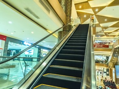 Parlak ışıklar ve çeşitli mağazalarla çevrili, yükselen yürüyen bir yürüyen merdivene sahip modern alışveriş merkezi içi, canlı ve yoğun bir perakende ortamı sergiliyor.