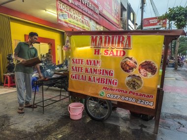 Street food vendor grilling satay at a roadside stall in Indonesia. The banner advertises chicken satay, goat satay, and goat curry. Contact number for orders is visible, Indonesia, 1 November 2024