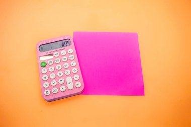 1234 sayısını gösteren pembe bir hesap makinesi turuncu bir arka planda parlak pembe bir kare kağıdın yanına yerleştirilir. Canlı renkler görüntüyü görsel olarak çekici kılıyor