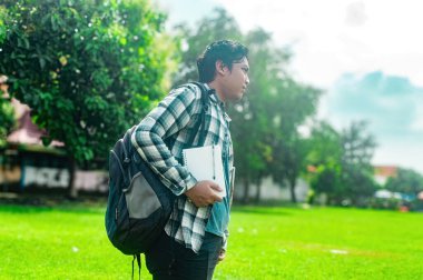 Sırt çantası ve not defteri olan bir öğrenci yeşil bir parkta ekose gömlek giyerek duruyor. Arka planda ağaçlar ve açık bir gökyüzü var. Bu da açık havada huzurlu bir çalışma ortamı olduğunu gösteriyor.