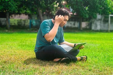 Parktaki çimlerin üzerinde dizüstü bilgisayar kullanan biri. Bireyler günlük giysiler giyiyor ve çalışıyor ya da dışarıda çalışıyor gibi görünüyor, doğal ortamın tadını çıkarıyor.