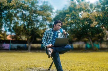 Parktaki katlanabilir bir sandalyede oturan biri elinde mavi bir kupayla dizüstü bilgisayarla çalışıyor. Arka planda yemyeşil ağaçlar ve güneşli bir gökyüzü var, sakin bir açık hava çalışma alanı oluşturuyor.