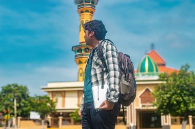 Sırt çantası ve not defteri olan bir öğrenci, uzun bir minare ve kubbeyle bir caminin önünde duruyor. Eğitimi ve kültürü simgeliyor.