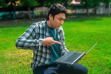 Parkta çimenlerde oturan, dizüstü bilgisayarı işaret eden, ekose gömlek ve mavi tişört giyen, mahremiyet için bulanık yüzlü biri.