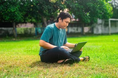 Çimenli bir arazide dizüstü bilgisayarla çalışan bir adam. Barışçıl açık hava manzarası ağaçlar ve güneş ışığını içeriyor, modern, uzak bir iş yaşam tarzını vurguluyor..