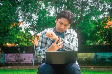 Kucağında dizüstü bilgisayarla dışarıda oturan bir adam, akıllı saatleri kontrol ediyor. Arka planda yemyeşil ağaçlar ve renkli bir duvar var. Bu da rahat ve verimli bir ortam olduğunu gösteriyor.